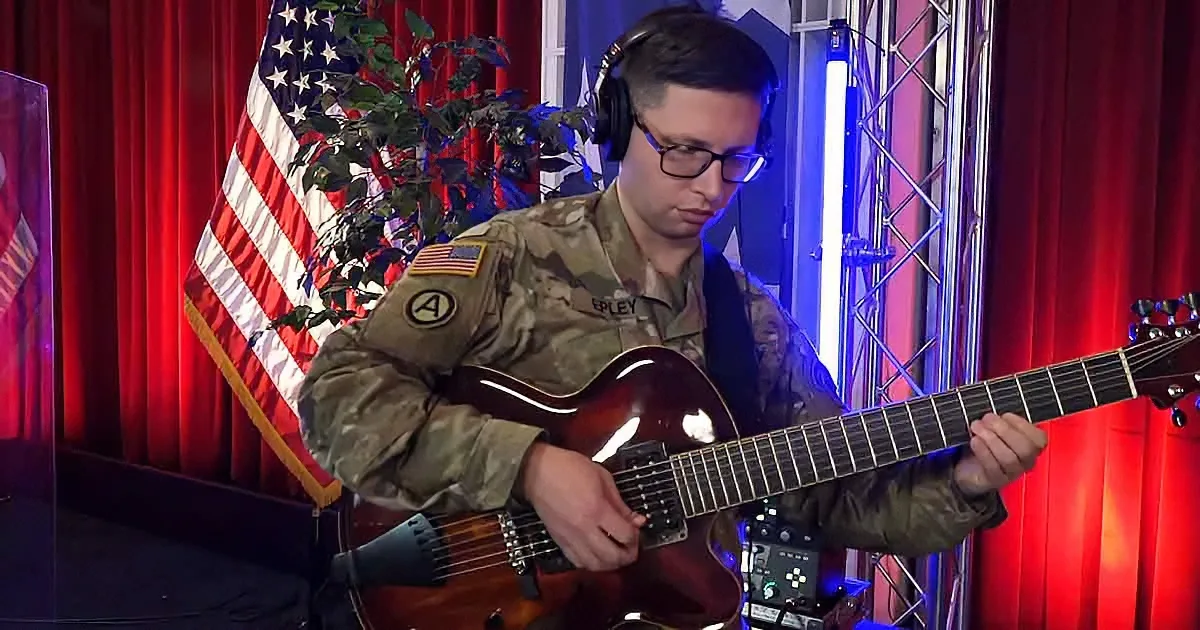 A soldier in uniform plays a guitar on stage with an American flag and red curtains in the background.