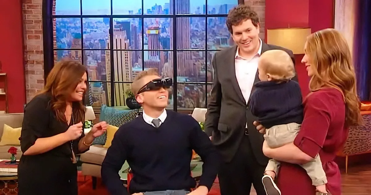 A man in a dark sweater and sunglasses sits, smiling, with a woman in a red dress holding a baby and another woman in...
