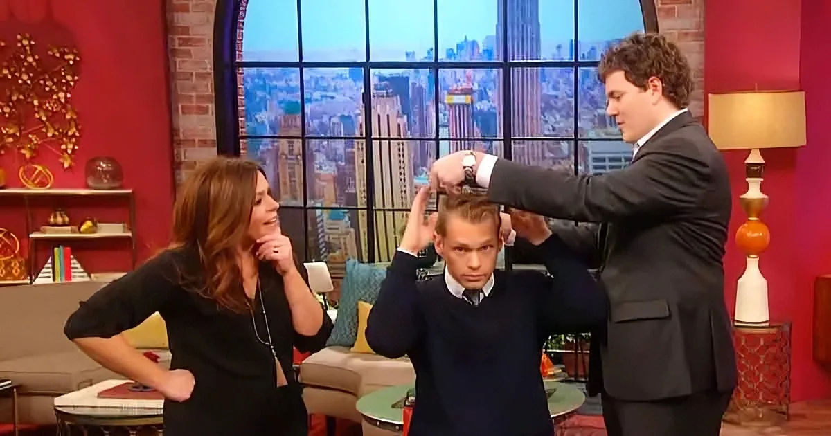 A man in a suit adjusts another man's hair on a talk show set with a cityscape backdrop.