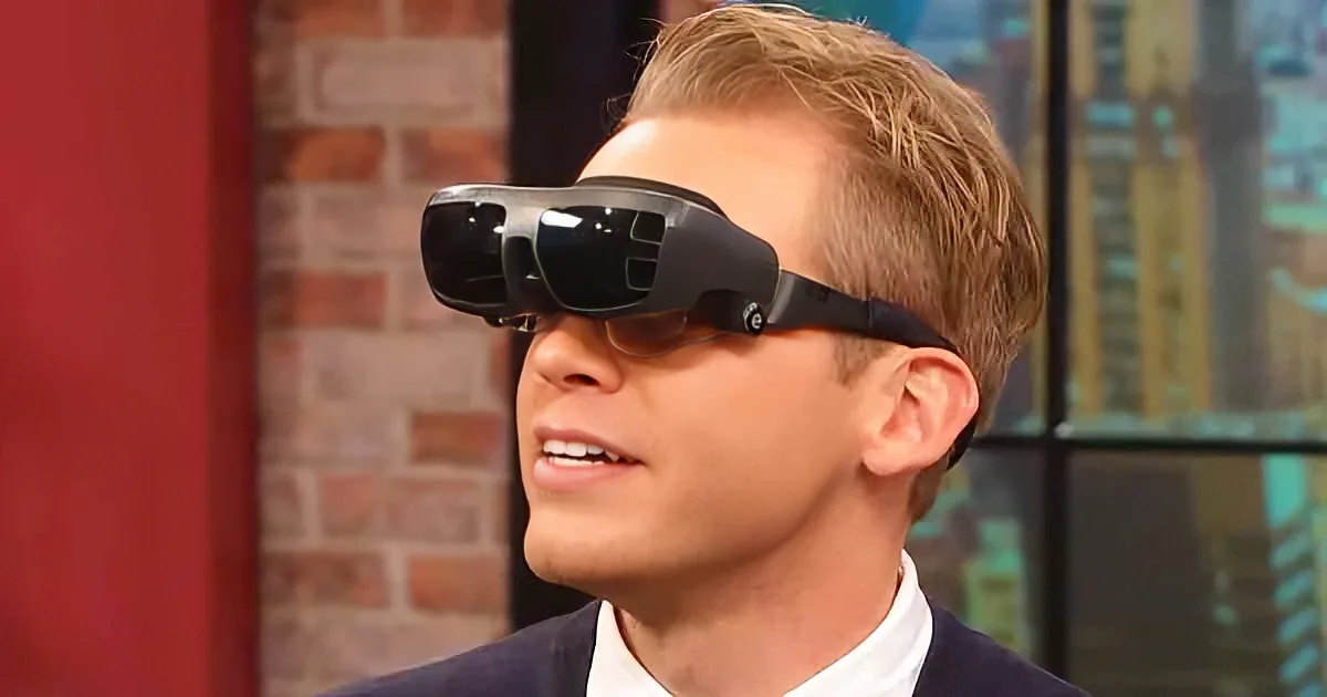 A man wearing a suit and a black VR headset, looking up, with a brick wall and cityscape in the background.