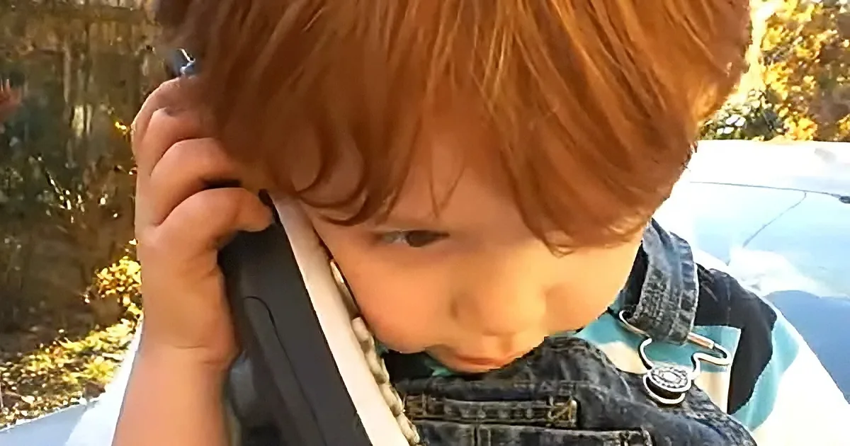 A child with red hair wearing a denim jacket is holding a phone to their ear.