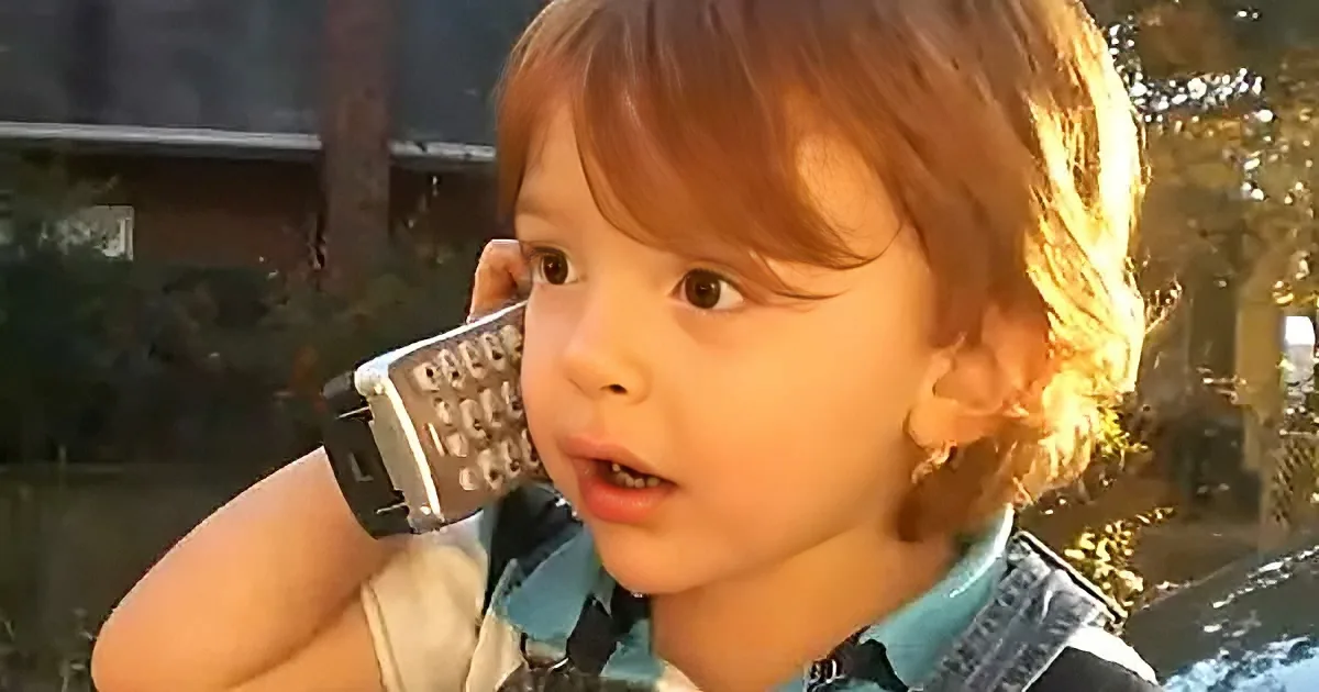 A young child with light brown hair holds a small, old-fashioned phone to their ear, appearing to listen intently.