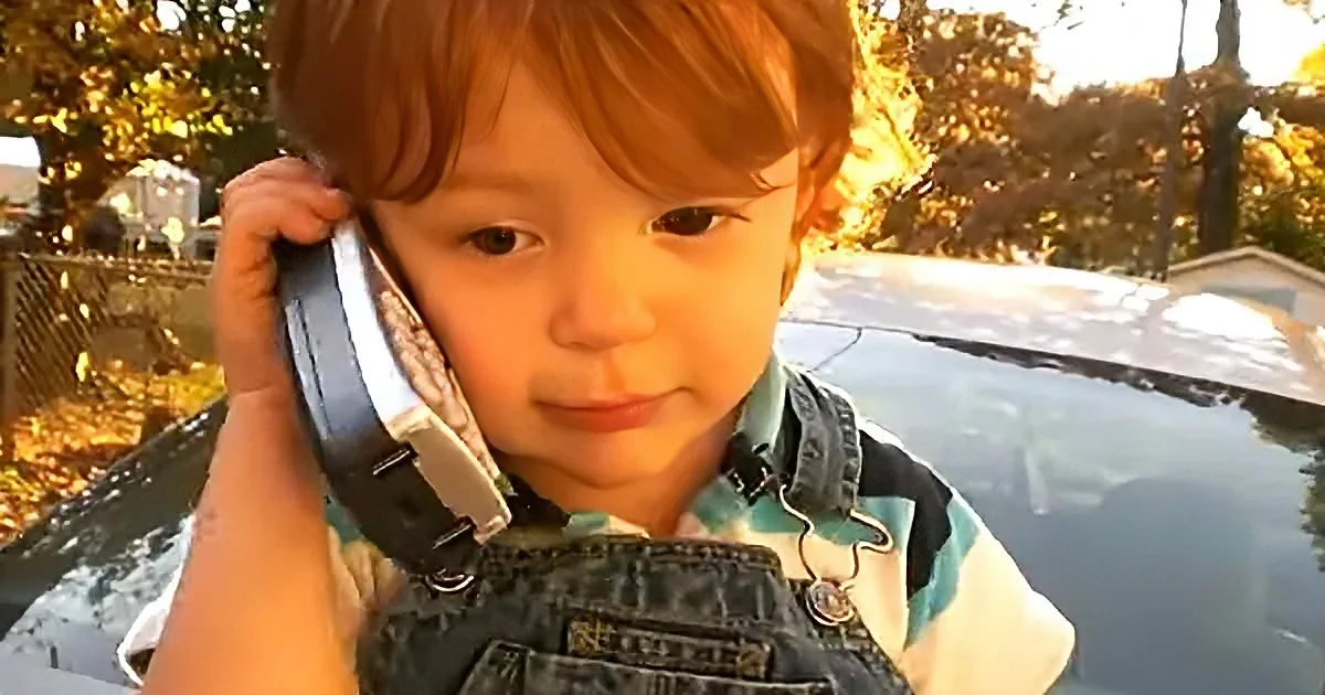 A young child with red hair holds a large, old-fashioned phone to their ear, wearing denim overalls and a striped shirt.
