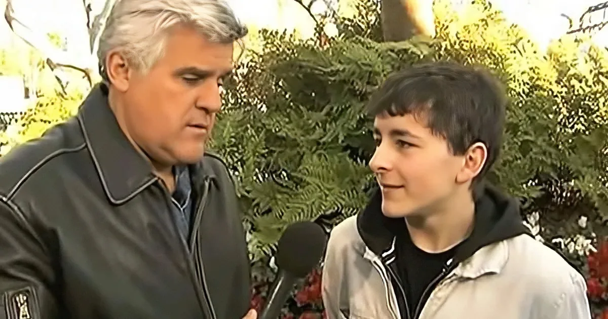 A man in a black jacket holds a microphone, interviewing a boy in a light jacket outdoors.