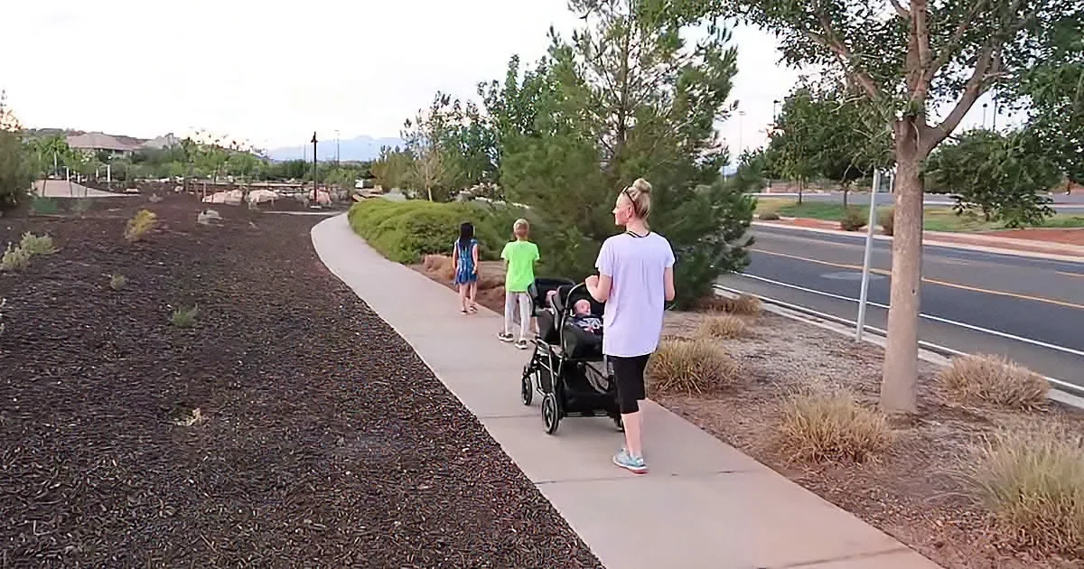 A woman pushes a stroller on a sidewalk near a road, with two children walking ahead.