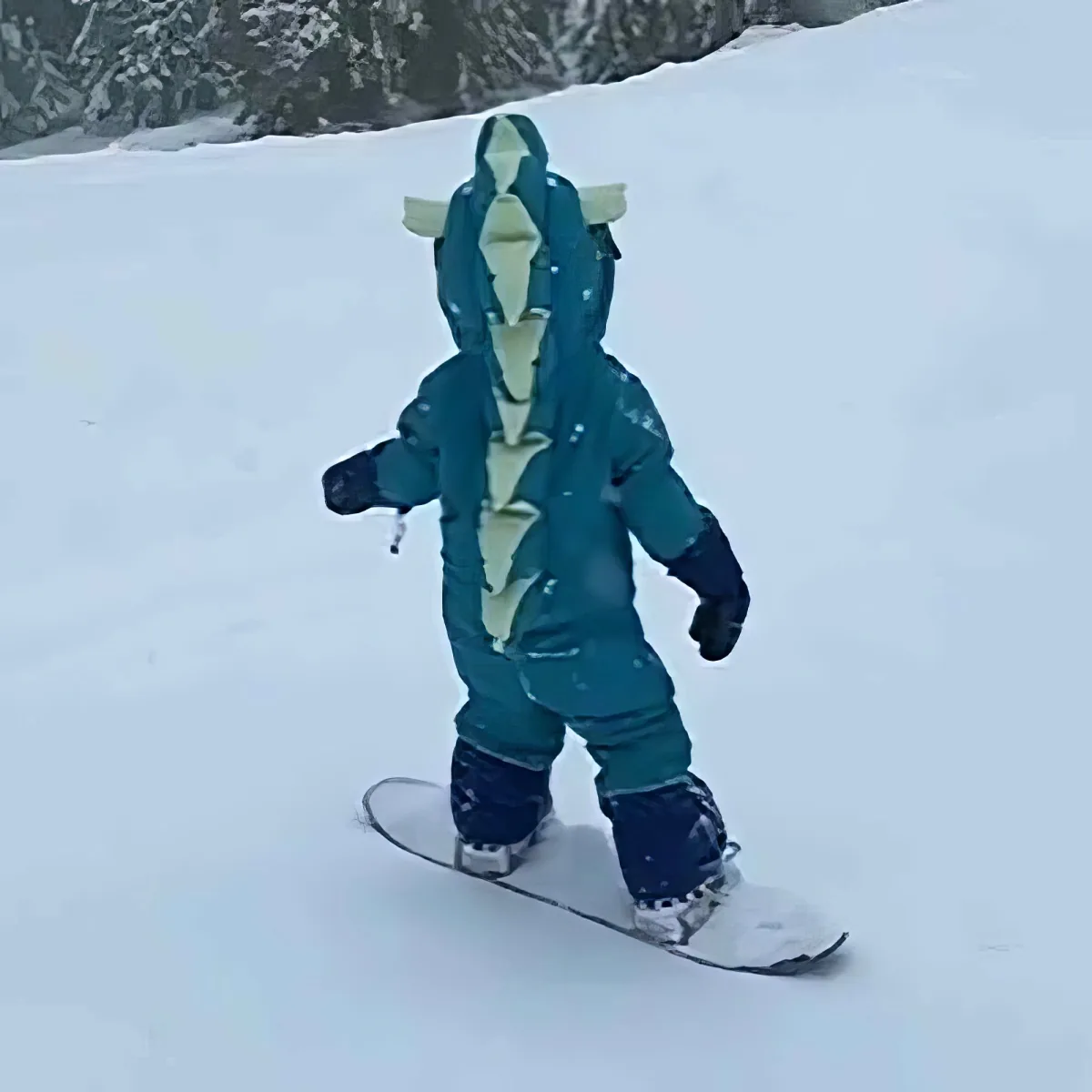 A child in a teal snowsuit with a shark design is snowboarding on a snowy slope.