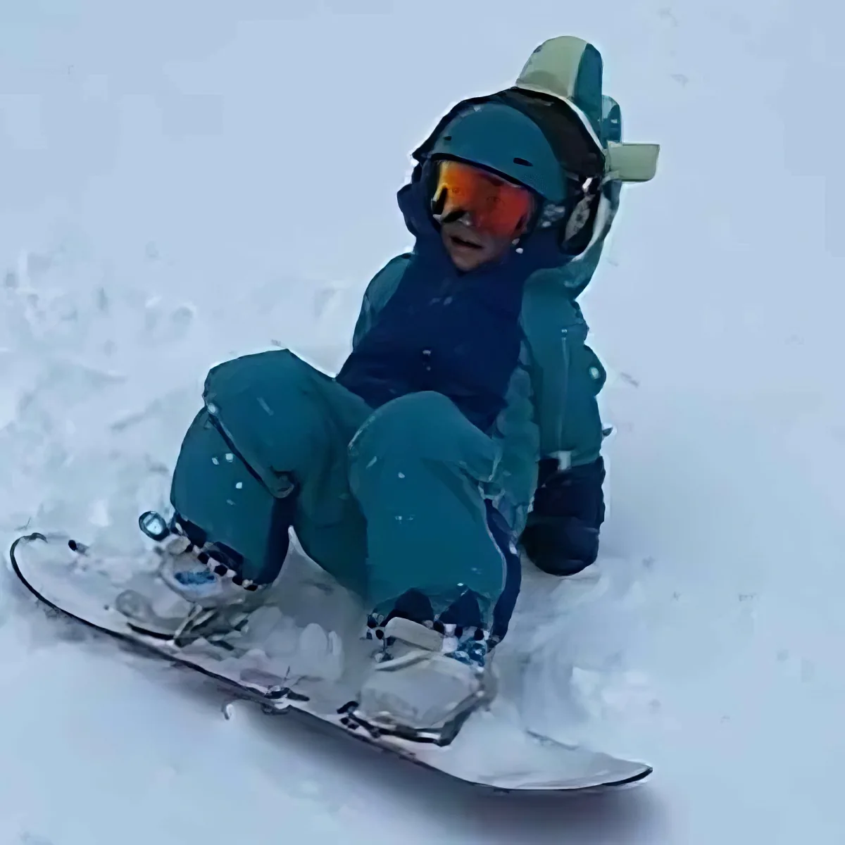 A person in teal snow gear sits on a snowboard, surrounded by snow, wearing a helmet and goggles.