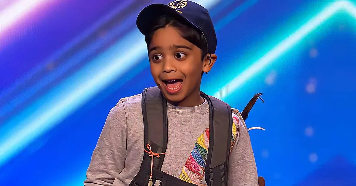 A young boy wearing a cap, backpack, and grey sweater with colorful stripes on the sleeve is seated on a stage with a...
