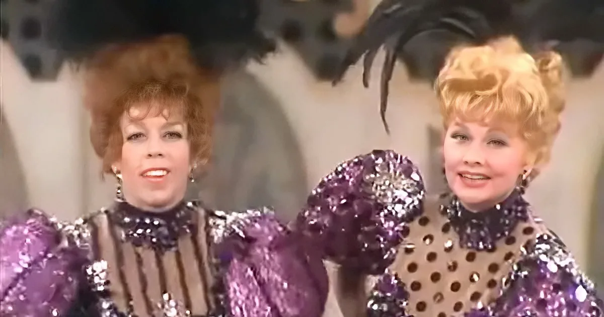 Two women in sparkling purple costumes with polka dots and feathered hats are smiling.