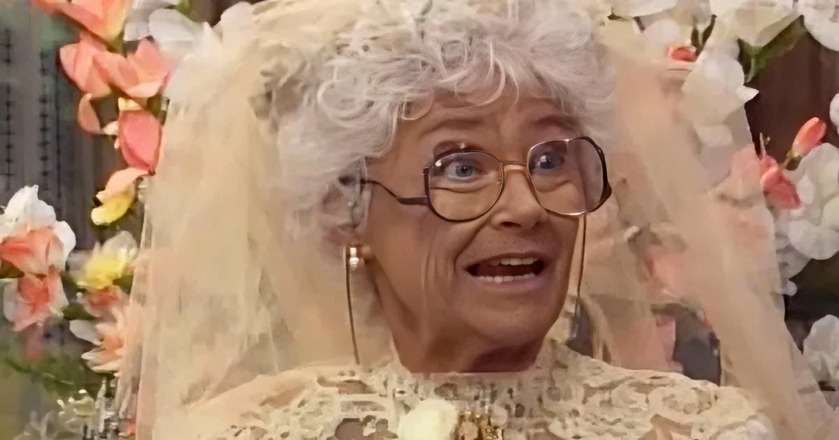 An elderly woman with white hair, wearing glasses and a floral dress, surrounded by flowers.