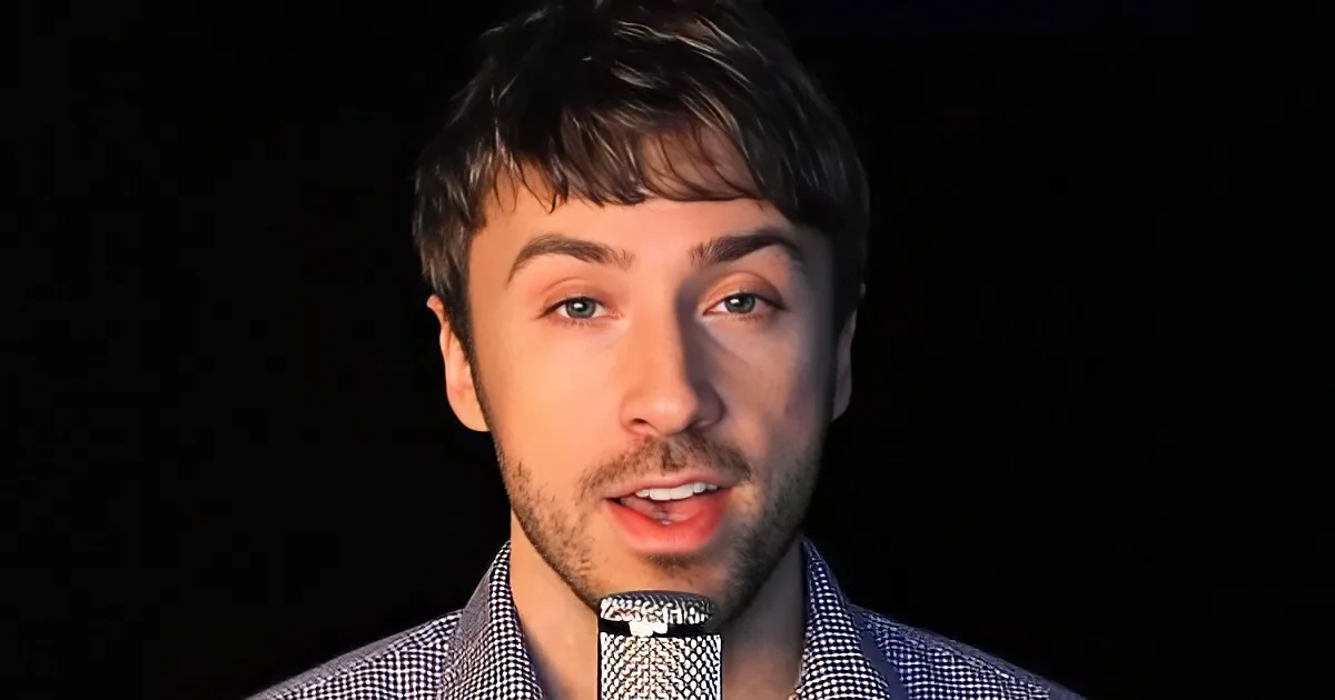 A man with short brown hair and a checkered shirt speaks into a microphone against a dark background.