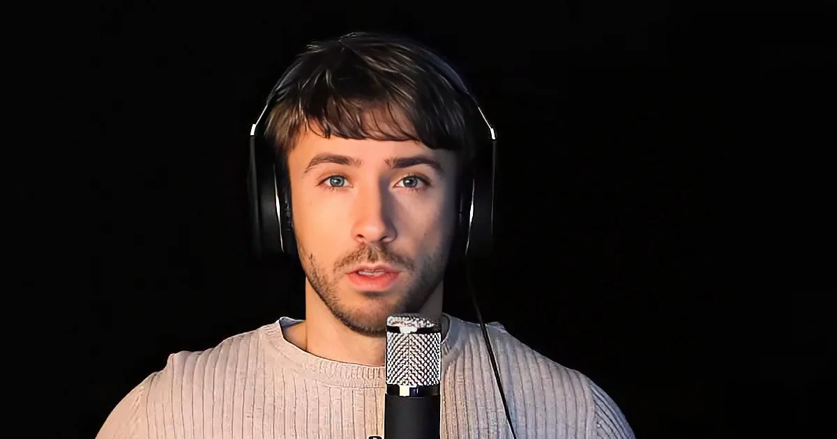 A person wearing headphones and a sweater speaks into a microphone against a dark background.