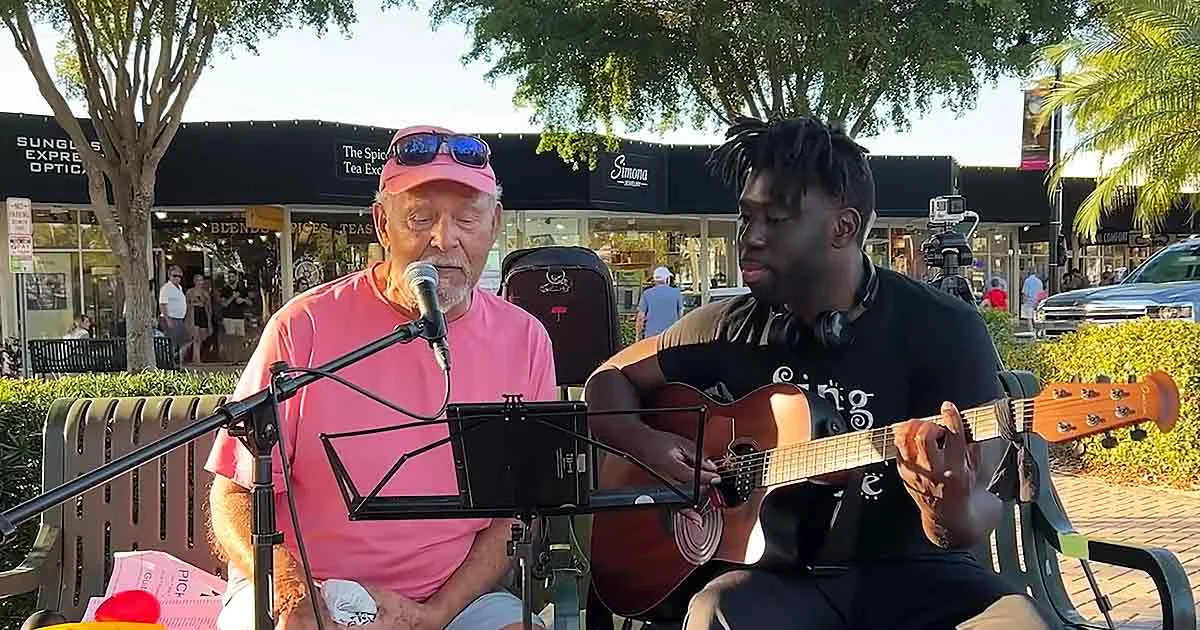 An older man in pink sings into a microphone while a younger man plays guitar on a bench.