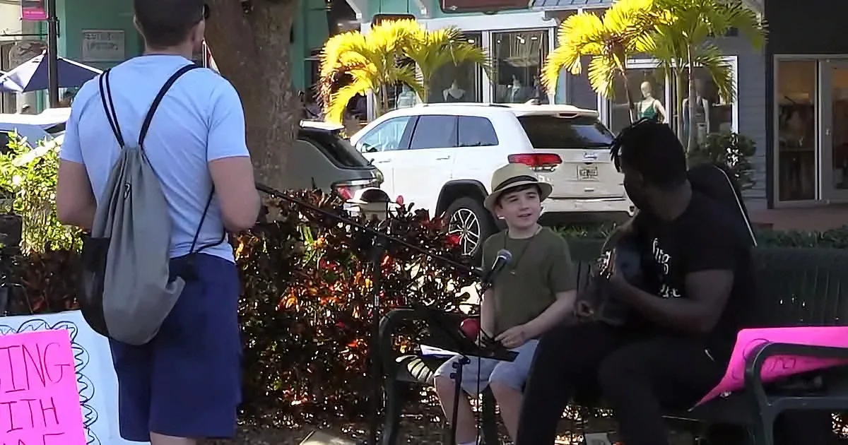 A young boy in a hat sings into a microphone while a man plays guitar in a public setting.