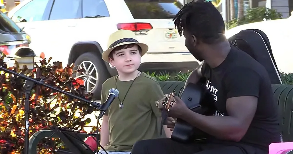 A young boy in a hat sits next to a man playing guitar, both in front of a microphone and a parked car.