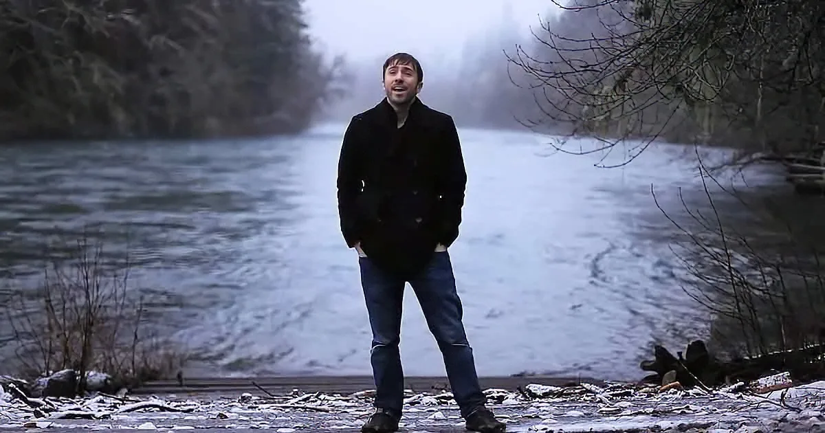 A man in a black coat stands on a snowy riverbank, smiling, with a misty forest backdrop.