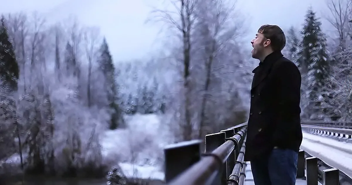 A man in a black coat stands on a bridge, looking up at a snowy forest.