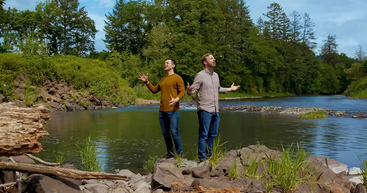 Two men stand on rocks by a river, arms outstretched, surrounded by greenery and trees.