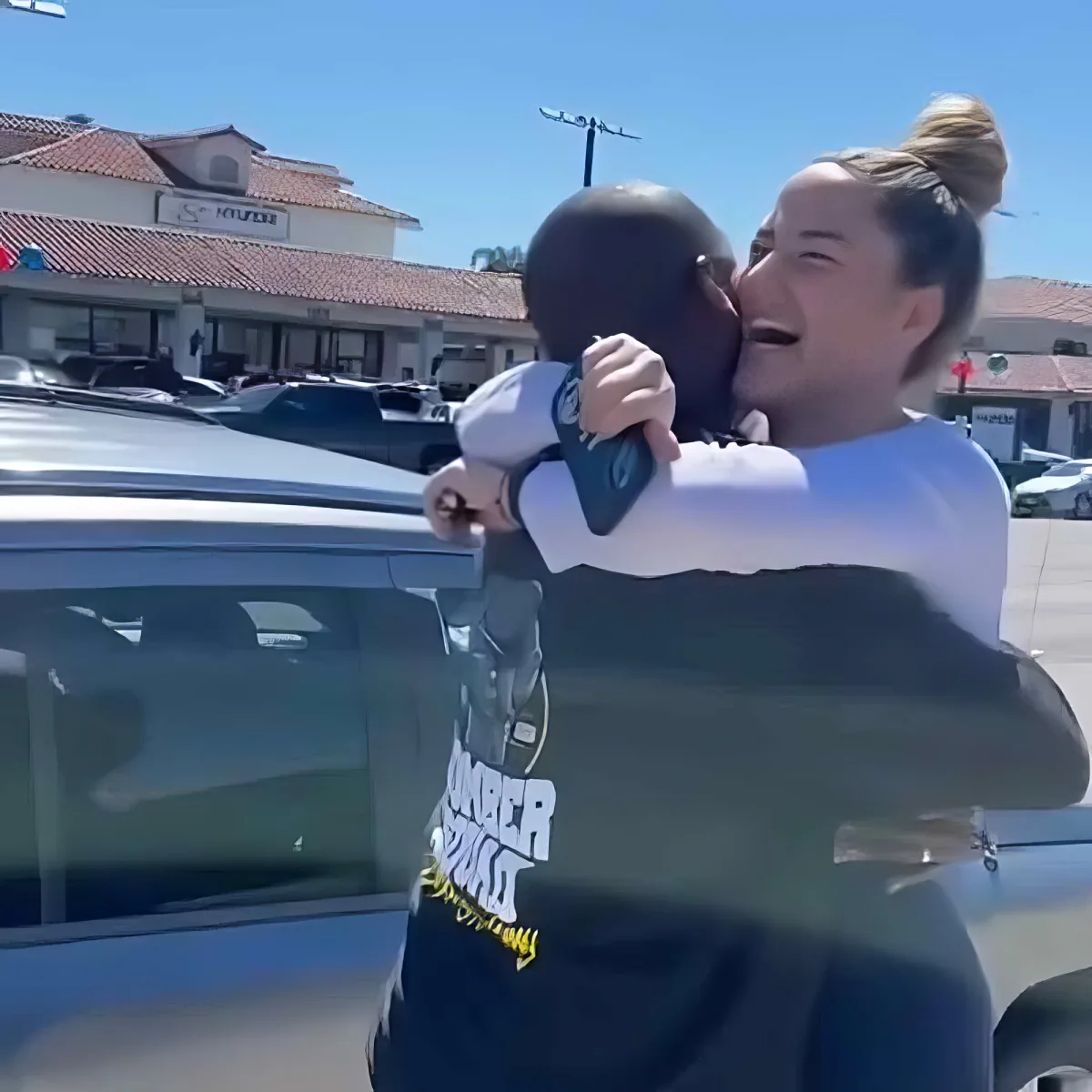 A woman hugs a child in a parking lot, both smiling, with a car and buildings in the background.