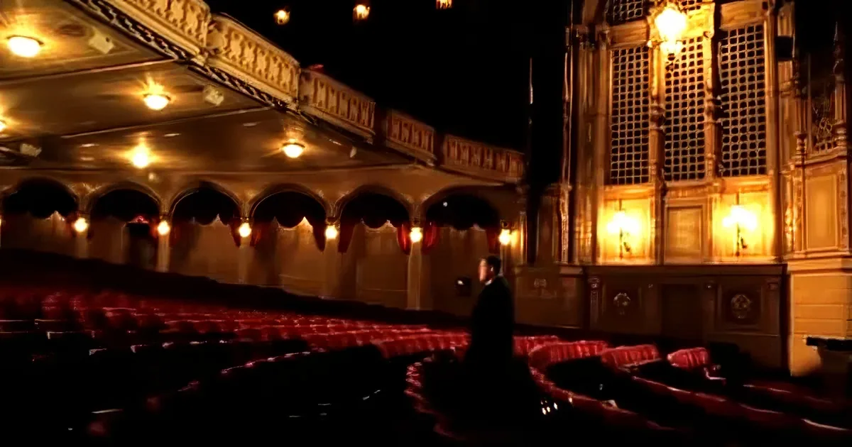 A man in a suit walks through an ornate theater with red seats and warm lighting.