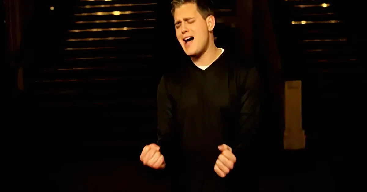 A person in a black shirt stands with clenched fists, mouth open, against a dark background with vertical light streaks.