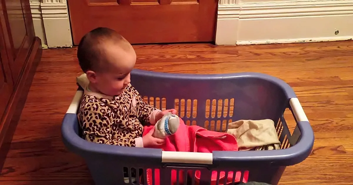 A baby in a leopard print outfit sits in a laundry basket, holding a stuffed animal.
