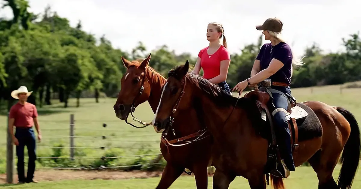 Two women ride horses in a field, guided by a man in a cowboy hat.