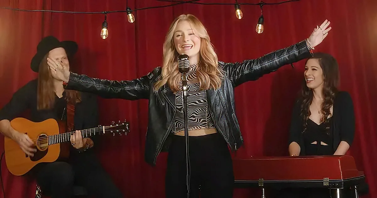 A woman in a black leather jacket sings into a microphone, arms outstretched, with a guitarist and keyboardist behind her.