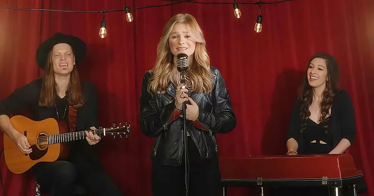 A woman in a leather jacket sings into a microphone, accompanied by a guitarist and a pianist.