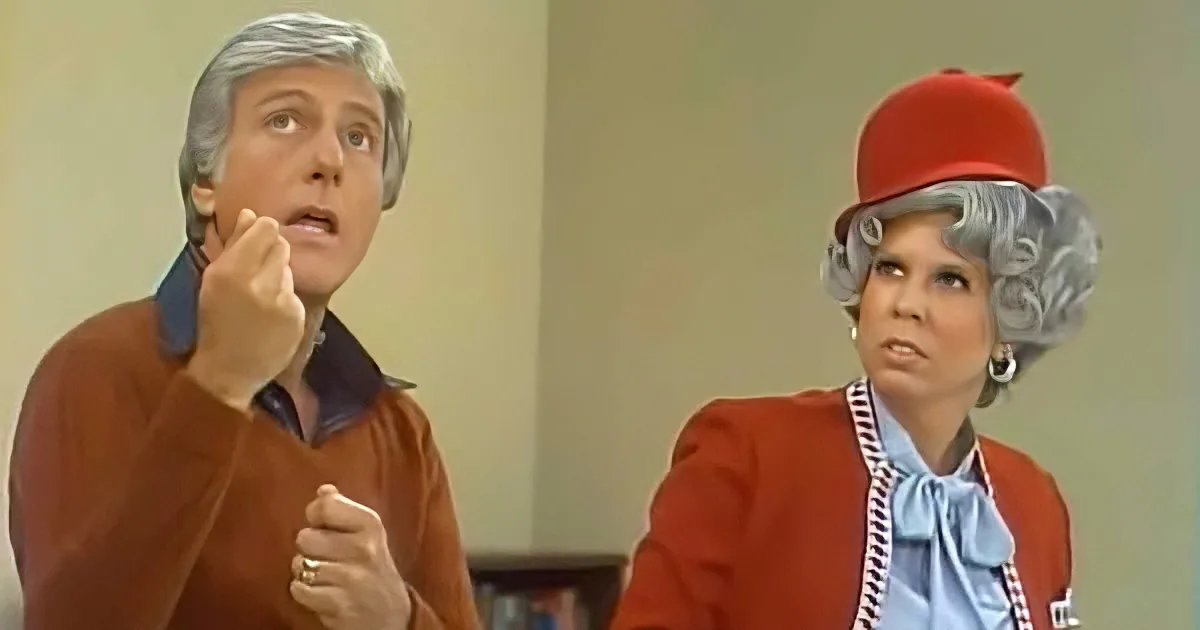 A man in a brown sweater gestures while a woman in a red outfit and hat looks concerned.
