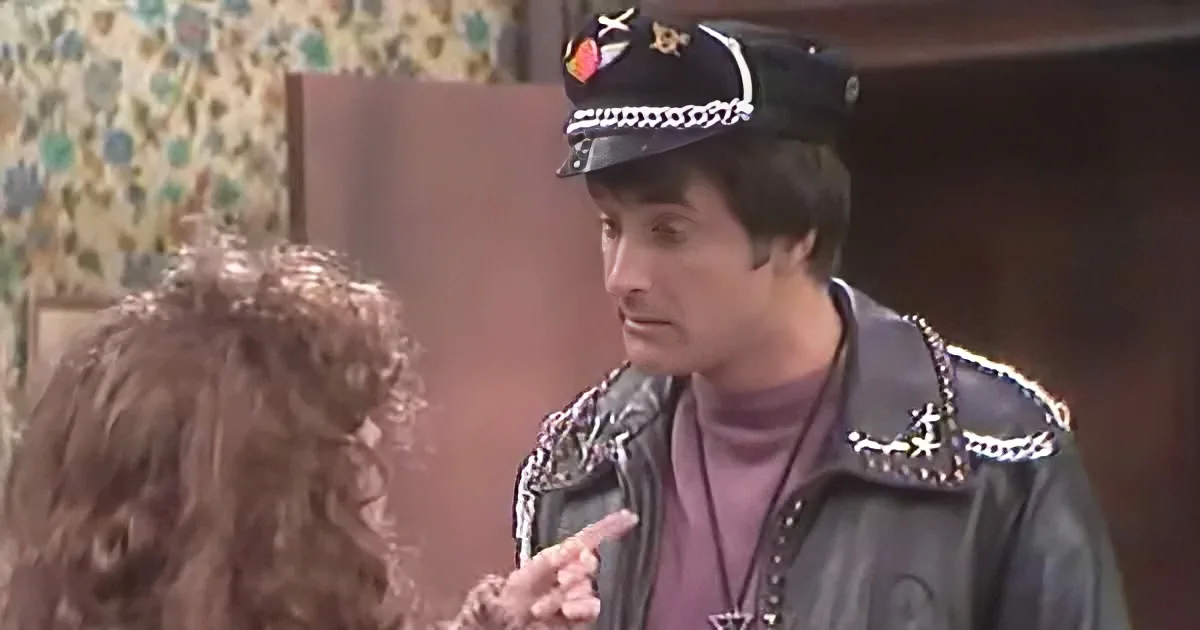 A man in a leather jacket and police hat is talking to a woman with curly hair.