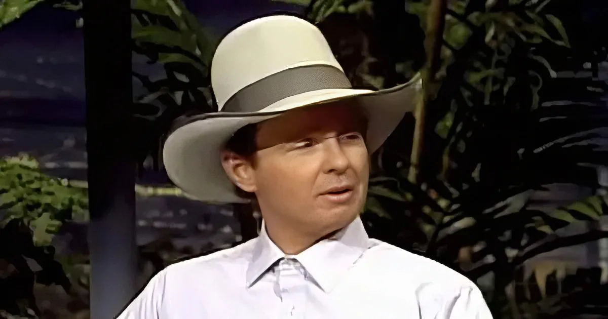 A man in a white shirt and wide-brimmed hat stands against a backdrop of tropical foliage.