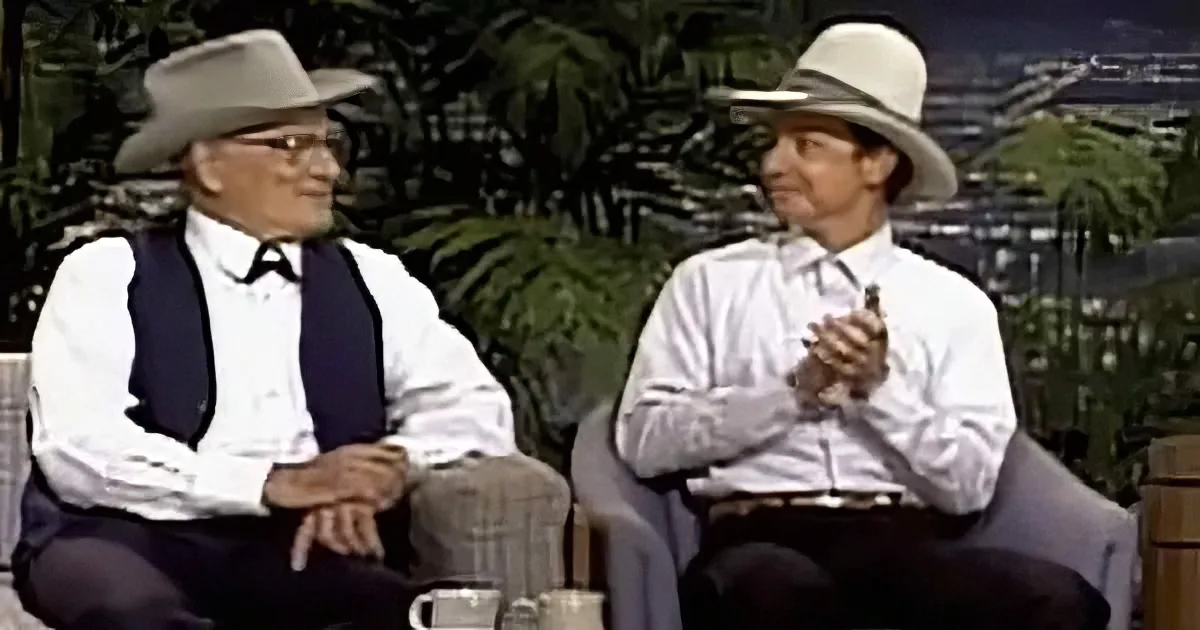 Two men in cowboy hats and attire sit on a couch, holding cups, with a tropical backdrop.