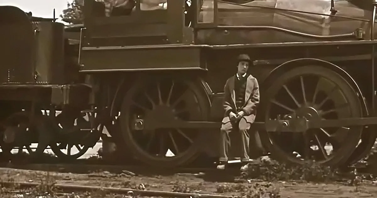 A man in a bowler hat sits on a train wheel, surrounded by sepia-toned railway scenery.