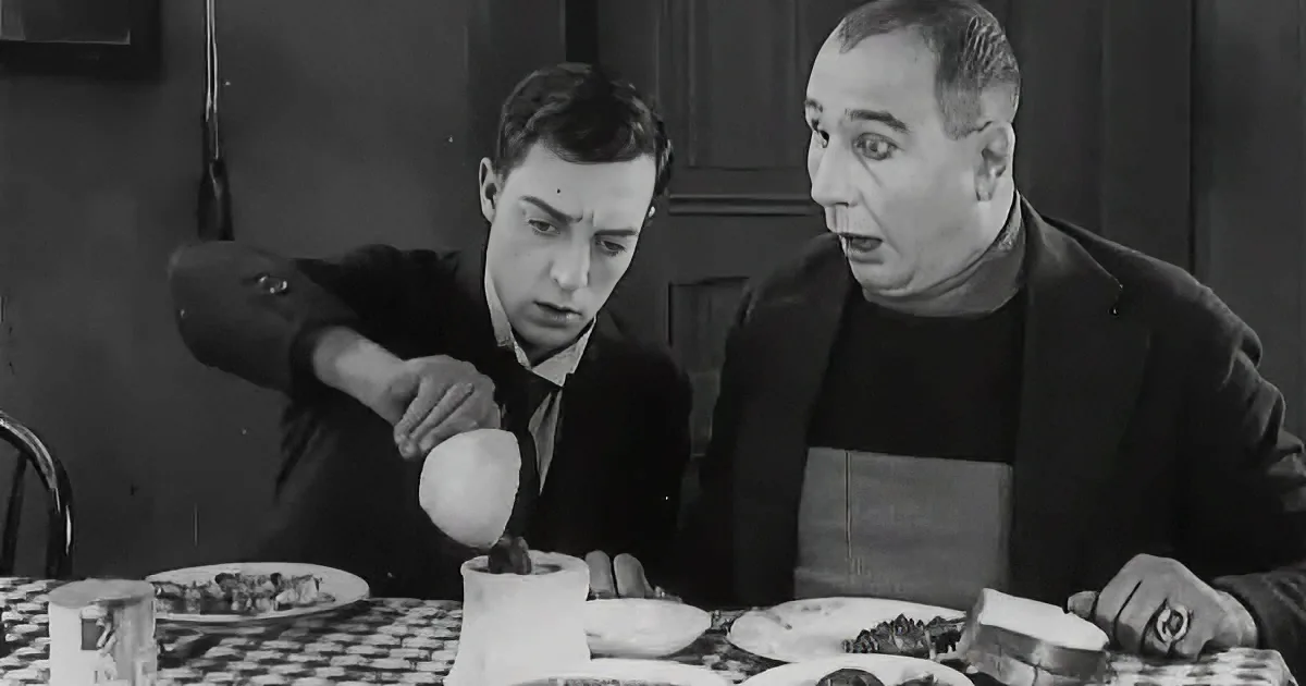 A man in a suit pours something into a cup while another man looks on, both seated at a table with plates of food.