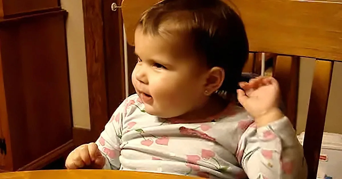 A baby in a cherry-patterned shirt sits in a wooden chair, holding a small object near their mouth.