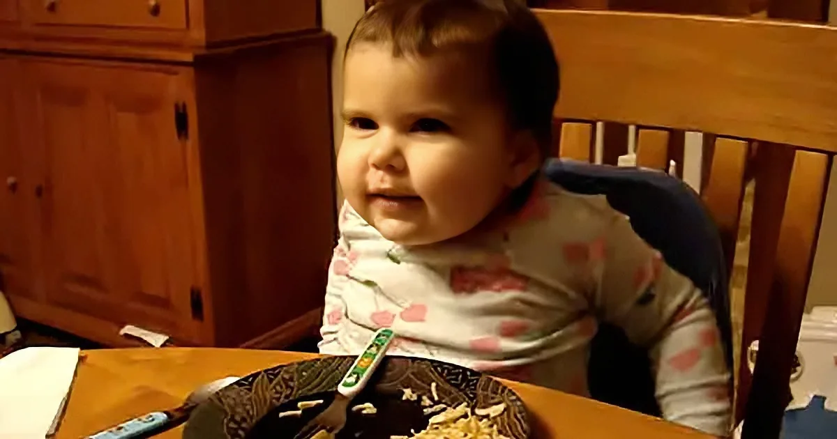 A baby sits in a high chair at a table, wearing a bib and looking to the side.