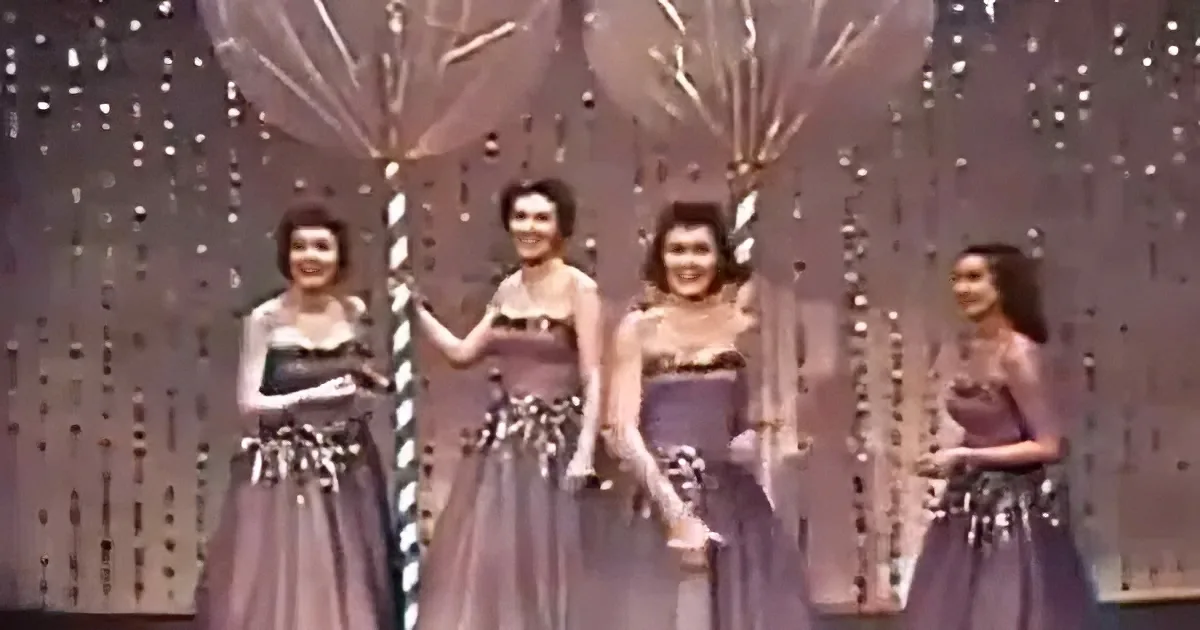 Four women in matching dresses stand on a stage with decorative lights behind them.