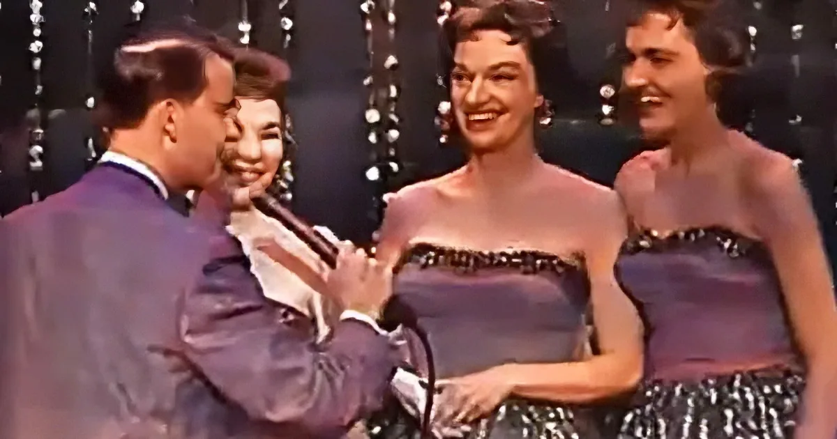 A man in a suit holds a microphone, smiling at three women in matching dresses.