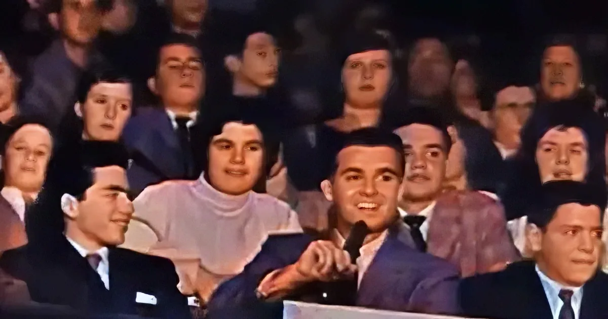 A man in a suit speaks into a microphone while an audience watches attentively.