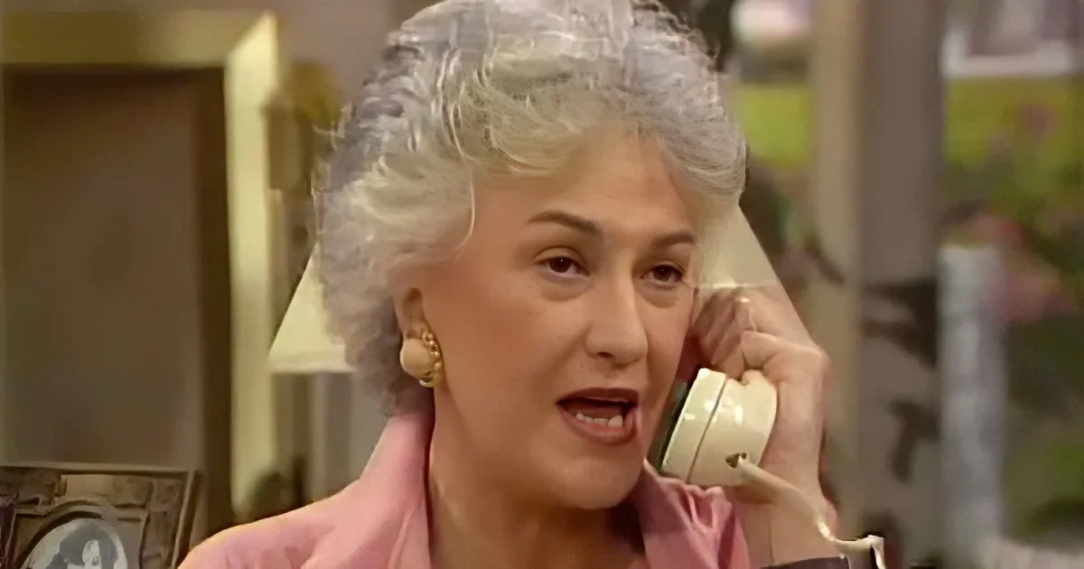 An older woman with white hair, wearing a pink jacket, holds a white telephone to her ear, appearing to be engaged in a...