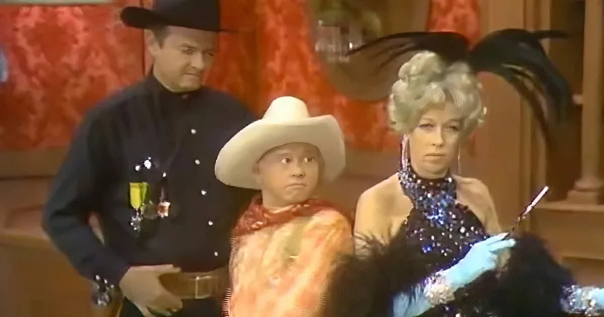 A man in a cowboy hat and colorful outfit sits next to a woman in a sparkly dress, both looking at something off-screen.