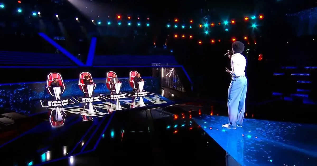 A performer stands on stage, facing judges seated in red chairs under colorful stage lights.