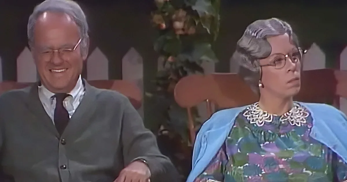 A man in a green sweater and tie smiles, while a woman in a floral dress and blue shawl looks concerned.