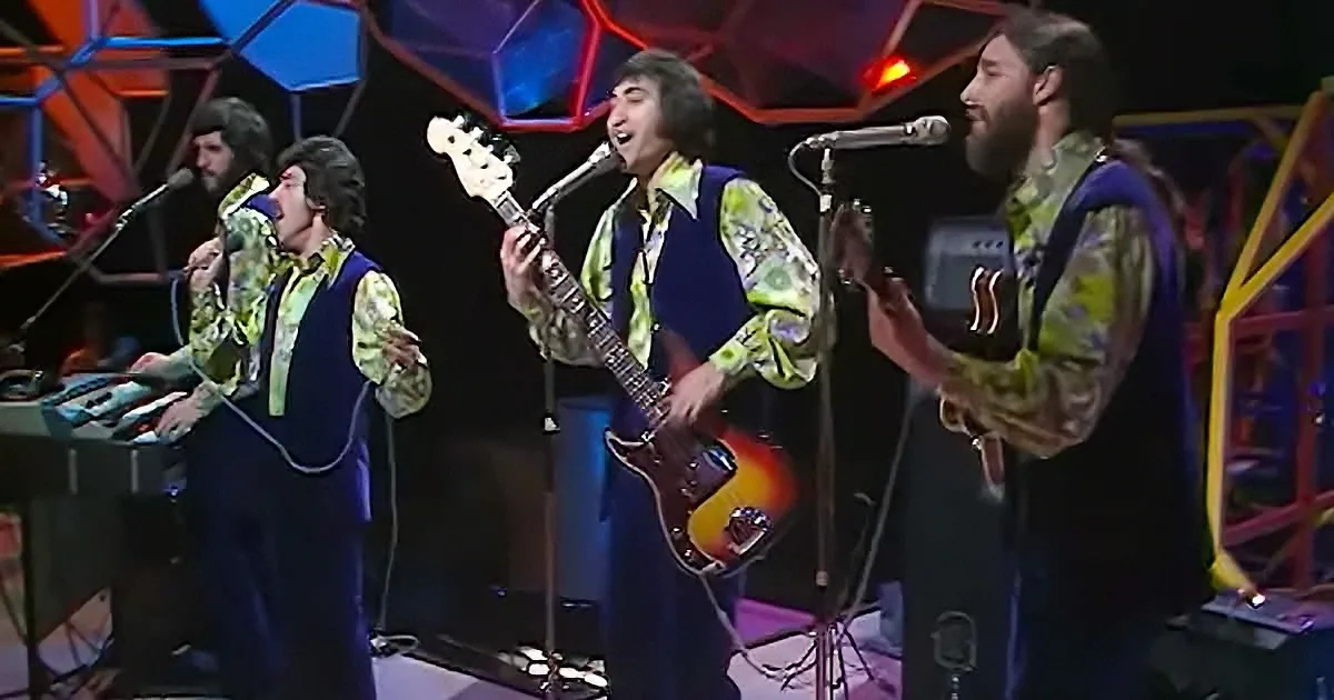 A band in vibrant 70s attire performs on stage with guitars and keyboards, surrounded by colorful geometric lights.