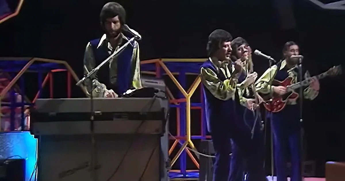 A man plays a keyboard while three others sing and play guitar on stage.