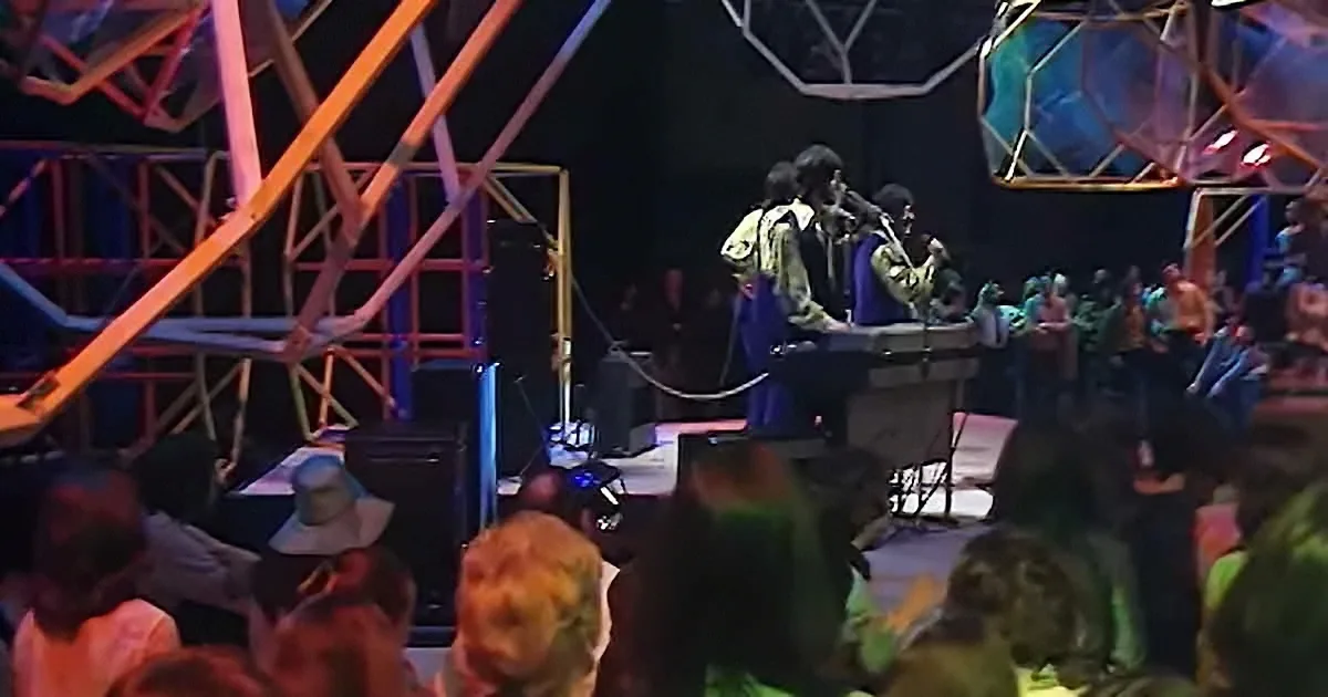 Musicians perform on stage with colorful lighting, surrounded by an audience.