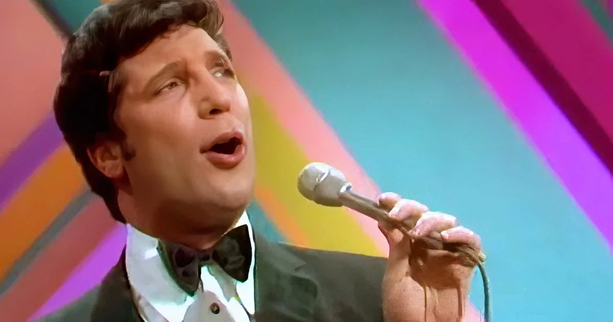A man in a suit and bow tie sings into a microphone against a colorful background.