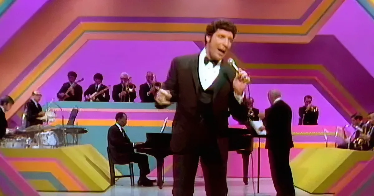 A man in a tuxedo sings into a microphone on a colorful stage with a band in the background.