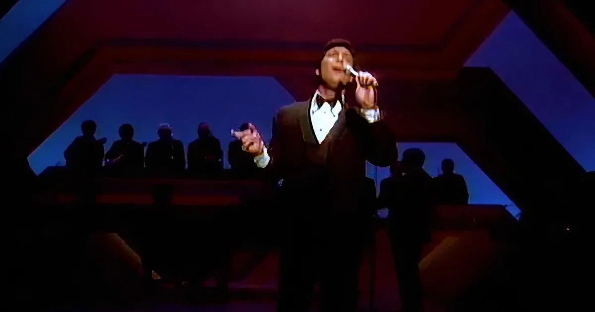A man in a tuxedo sings into a microphone on stage, with silhouetted figures in the background.